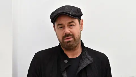 BBC Danny Dyer stood in front of a white wall. He is wearing a black top, jacket and hat.