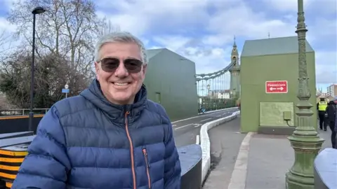 Chris wears a blue puffer jacket, has white short hair and is stood by the entrance to Hammersmith bridge.