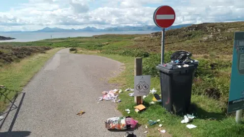 BBC A wheelie bin overflowing with litter, next to a "No Entry" sign. It sits next to single track road which leads off into the distance. In the background is a stretch of water and a picturesque, mountainous landscape. 