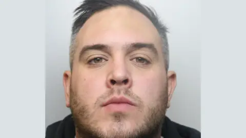 A police custody image of Mitchell Curtis. He has short grey hair and is wearing a black hooded jumper.