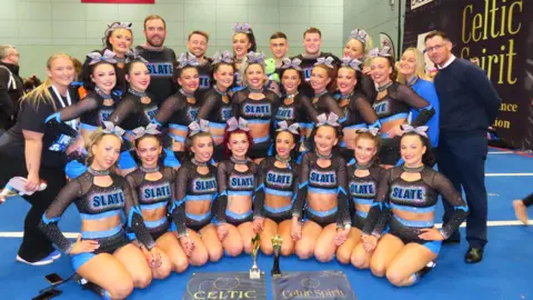 Strikeforce Cheer A number of cheerleaders and their coaches are standing side-by-side in a team photo. They are all dressed in their black and blue cheer uniforms with the word Slate written across their chests. The female members are all wearing ribbons in their hair. 