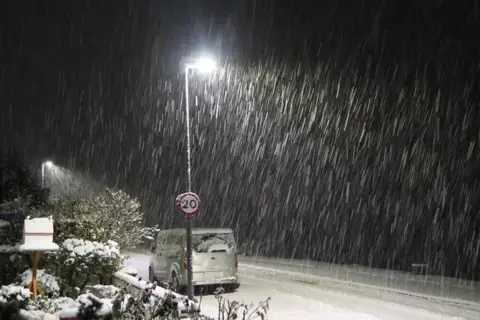 BBC Weather Watchers/Ingrid Snow falls in the dark hours of the morning in Lochcarron in the Highlands. A snow covered garden can be seen as well as a car and a 20mph road sign.