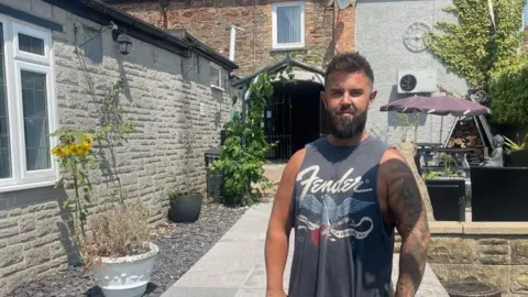 Joshua Collins, a young man with dark hair and a big beard, is wearing a vest in the garden of a pub.