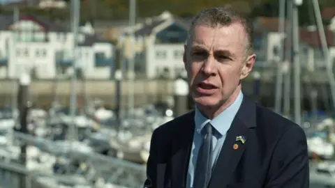 Adam Price looking just to the left of the camera and speaking to a reporter. He is standing in front of a seaside setting with boats and buildings behind him.
