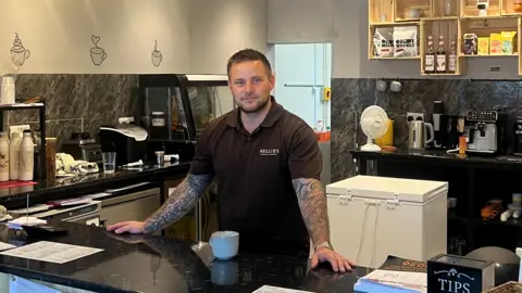 A man with tattoos on both arms stands over a counter at a coffee shop.