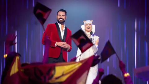 Rylan and his co-host of the Interstellar Song Contest in Doctor Who stand on stage as people wave flags in the air