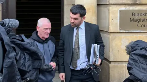 Ollie Samuels/BBC News A middle-aged man with thinning white hair and a younger man in a suit walk out of a sandstone court building. 