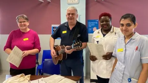 Shrewsbury and Telford Hospital NHS Trust A man with grey hair is holding a guitar. Around him are three women who work at the hospital, one dressed in light bur scrubs, the other in white uniform, and the other in a pink jumper.