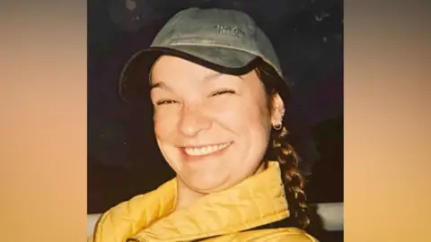 Clare Dupree, smiling, and wearing a yellow coat and green baseball cap. She has brown plaited hair 