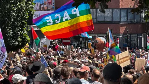A group of people holding a variety of flags. There is a large multi-coloured flag, with the word "pace" printed on the flag.