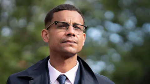 PA Photo of Daniel Kebede. He has short hair and is wearing glasses and a shirt, tie and smart navy blue jacket.