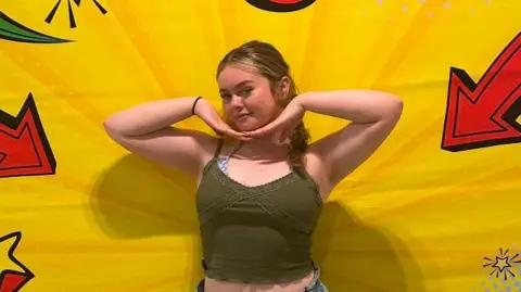 Grace A young woman poses centered against a vibrant, pop-art style backdrop with bright yellow burst patterns and comic-book style red and black arrows or stars pointing inward.