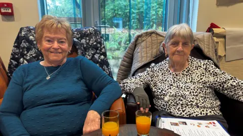 A photo of a lady with short, blonde hair and blue jumper sat in a chair next to a lady with short grey hair and a black and white speckled jumper.
