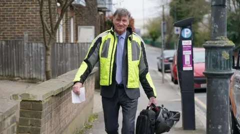 Lord Brocket carrying his moped helmet, wearing a hi-viz jacket