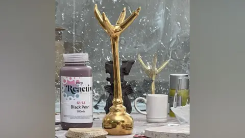 Simon Balch A gold plated spherical egg cup holder in the shape of an upside down chicken claw sits on a table surrounded by other items like a mug and some paint. 