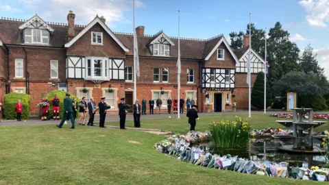 A line of people dressed in uniform in front of a pond with a ring of bunches of flowers round it.