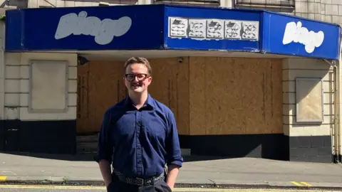 BBC The former Mecca Bingo site boarded up. A man wearing glasses and a blue shirt stands smiling in front.