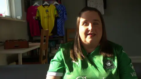 A woman wearing a green Plymouth Argyle football shirt sitting. Behind her, football jerseys in different colours, including yellow and blue, hang on a rack. A mini-foosball table is visible in the background on top of a table, and sunlight streams into the room, partially shadowing her face.