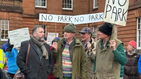 A group of people stand outside a sandstone building with signs reading "No Park"