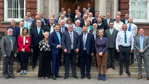 Reform UK A large group of smartly dressed people stand on the steps of a council building and smile towards the camera.