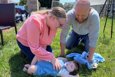 Family photo Amanda and Nick are kneeling on grass playing with Jack. Amanda has a pink long-sleeved top, wears glasses and has her fair hair tied back in a pony tail. Nick has a beard and is wearing a grey jumper and jeans. Jack is in a blue baby grow and has a feeding tube going up his nose.