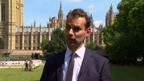 Josh Simons, who has brown hair, and is wearing a blue siut and white shirt, speaks to the camera outside Westminster