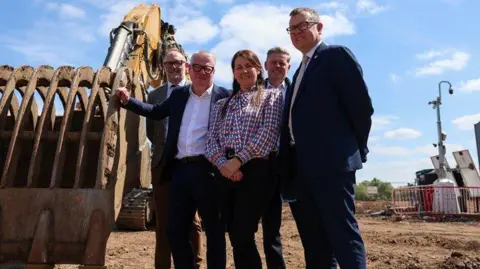 Four people stood in front a construction site with a large JCB digger 