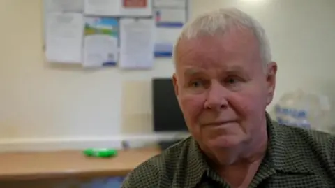 Mark is photographed indoors, in a room with beige walls and a pin board just behind his head. He's wearing a green collared shirt and has a sad expression on his face. 