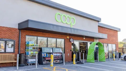 The exterior of a food store. There is green lettering which reads Coop and a large green archway in front of the store's entrance. There are display cabinets situated in front of large glass windows and a bench underneath a noticeboard to the left hand side of the image.