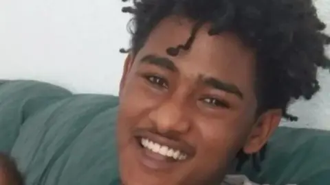 A close-up of a young man, with black curly, smiling towards the camera. He is sat on a green sofa.