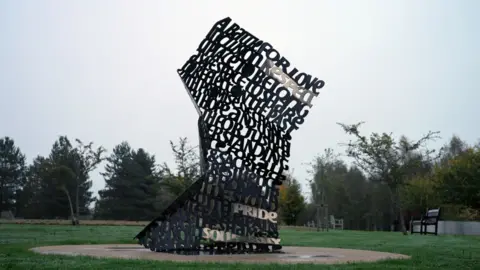 BBC / Ann Gannon A picture shows the monument stood in the National Memorial Arboretum, with grass around it and trees in the background. The monument itself is shaped like a large crumpled note made of metal words. You can make out "a battle for love" at the top, while words like "peace", "pride" and "belong" stand out further down.