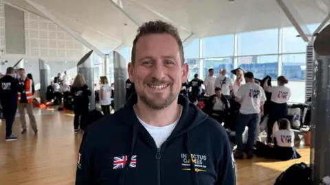 Scott Robertson is pictured smiling and wearing a blue hoodie with a union jack printed on the left hand side of the shirt. He has brown curly hair. He is inside Birmingham Airport