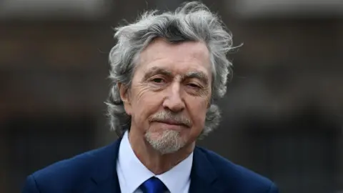 Mickey Brady, a man with wavy grey hair, a goatee beard and moustache, is wearing a navy suit with a white shirt and navy tie. The background is blurred. 
