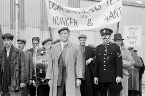 A group of twelve men in 1920s clothes are standing in a row in front of a corrugated iron building. They all look very serious and are clearly demonstrating. They have banners saying 'Down with the Means Test and Hunfer and Want'. One has a large drum. A policeman stands beside them.