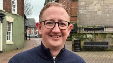 Charlie Stubbs/BBC News A man in a dark navy blue zip-up jacket with black framed glasses and short, brown curly hair. He is stood in a town square with a green building with a blue door and white windows to his left, and old grey brick building with black railings, a black litter bin and a bench, to his right
