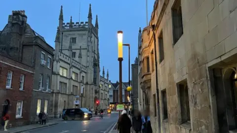 Steve Hubbard/BBC A lamp on a street in Cambridge at dusk. It is tall and thin and lit up. There are historical buildings on the street and pedestrians walking on the pavements. There is a van and a cyclist in the road