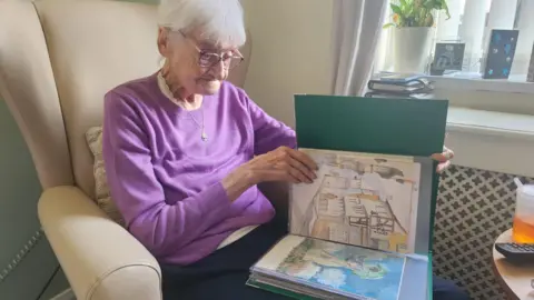 Provided Patricia, wearing a purple jumper, looking through her portfolio of paintings.