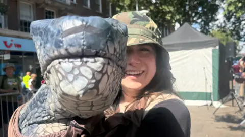 BBC / Sarah-May Buccieri The photo shows a woman smiling at the camera. She is wearing a camouflage hat and is holding up a dinosaur puppet. 