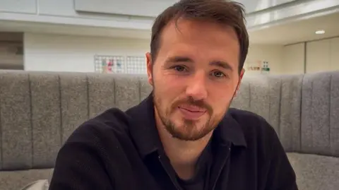 Hollie Donnellan Dominic McGregor sits in on a grey couch and wears a black polo neck long sleeved jumper. He has a beard and has short brown hair