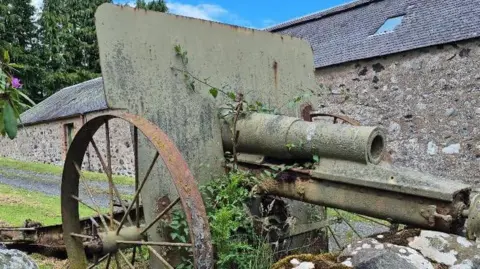 Great War Huts A close up of a rusty short-barrelled 10.5cm howitzer gun with its nose resting on a low stone wall. It has a large rectangular back plate between two wheels. The gun's barrel sticks out from the back panel. Beyond it is a low stone-built barn. 