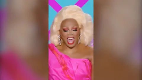 RuPaul in a bright pink outfit smiling widely