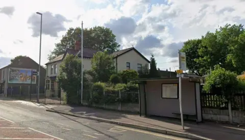 Carters Arms on Manchester Old Road Middleton is obscured by trees, a lampost and phone mast. There is a buys shelter and stop to its left.