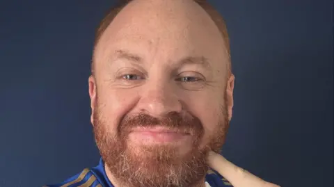 Image shows Eli's face. He is pointing at his beard which is fairly thick so far and is ginger. 