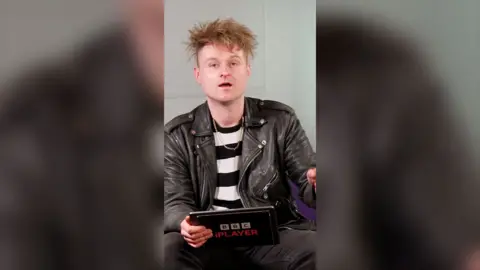 Sam Battle is sat wearing a black leather jacket over a black and white striped t shirt. He's holding a tablet.