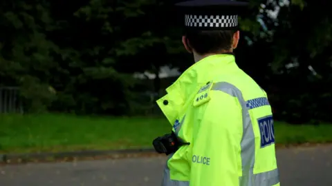 BBC Police officer in a hi-vis jacker with Police written on it. He is facing away from the camera and is also wearing a black police cap.