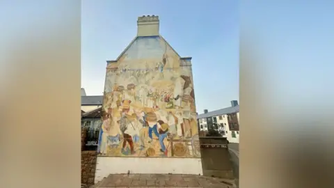 A 26 year old mural on the side of Jersey Museum in St Helier. It focuses on the history of the local area showing the bustling activity that took place between the military life of Fort Regent, the hotels and homes along Pier Road and the trade and commerce around the harbour.