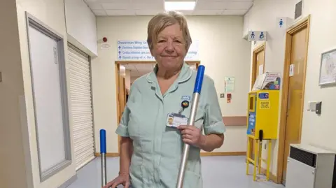 An older lady with short blonde hair in scrubs, standing in a hospital hallway, holding a mop handle.