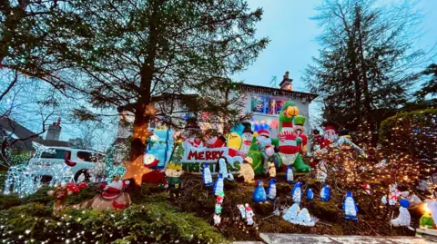 Caroline Eadie A Christmas display outside the front of a house in Giffnock. There are various Christmas figures on display including snowmen, Santa Claus and inflatable reindeer. A white house is visible in the background. A white car is parked in the drive.