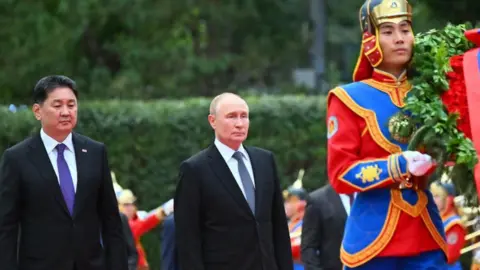 EPA Mongolië-president Ukhnaagiin Khurelsukh en de Russische leider Vladimir Poetin achter een Mongoolse man gekleed in traditionele rode en blauwe kleding. Achter hen zijn heggen.