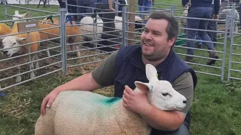A man with short brown hair, wearing a blue gilet and green T-shirt, has his arms around a sheep. He crouches in a livestock pen, surrounded by other sheep in metal pens.